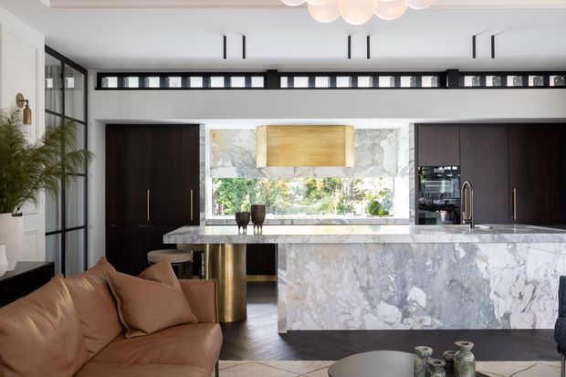 Open-plan kitchen viewed from an adjacent living area, featuring a large veined marble island and backsplash, dark timber cabinetry, integrated appliances, brass-toned pendant lighting, clerestory windows, and a tan leather sofa in the foreground. The space is bright, refined, and contemporary with a strong light marble and dark wood contrast.