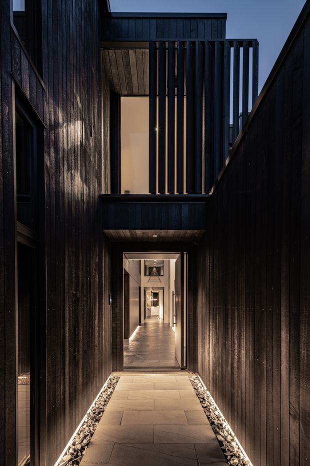 A dramatic narrow exterior passage leads toward a glazed entrance and illuminated interior corridor. The surrounding building is clad in dark vertical timber boards, with a projecting upper-level balcony or void screened by vertical timber battens. Large rectangular paving slabs form the walkway, bordered by strips of decorative pale stone and subtle linear lighting. The image is photographed at dusk with a moody, contemporary architectural character.