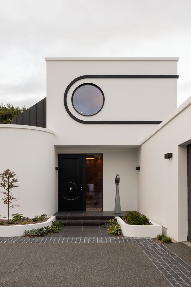 Front exterior view of a contemporary two-storey residence with smooth white rendered walls, a bold black curved geometric feature surrounding a circular window, flat roofline, black entry door, and a recessed entrance approached by dark tiled steps. White raised planter beds and dark paver bands define the landscaped forecourt.