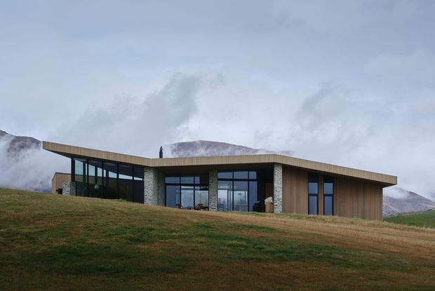 A low, single-storey contemporary house set on an open grassy hillside, with a broad shallow roof, extensive dark-framed glazing, vertical timber cladding, and pale stone masonry walls. Misty mountains rise behind the building, creating a remote alpine setting.