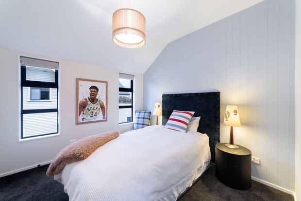Bright, compact bedroom with a white upholstered bed dressed in white bedding and striped accent cushions, a dark padded feature headboard, dark carpet, pale vertically panelled walls, and two narrow windows with blinds. Bedside tables, table lamps, a pendant light, and basketball artwork create a youthful contemporary feel.
