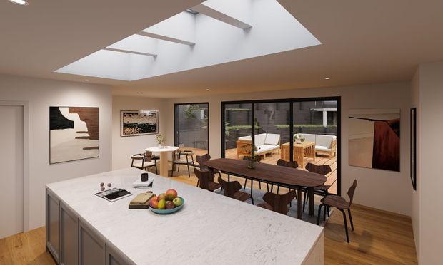 A bright, open-plan kitchen and dining area with a large pale stone island in the foreground, warm timber flooring, and dark-framed glazing opening to an outdoor courtyard. A long timber dining table sits beside the kitchen, while a large ceiling skylight brings in natural light. Neutral walls, contemporary cabinetry, artwork, and timber furnishings create a refined modern interior.