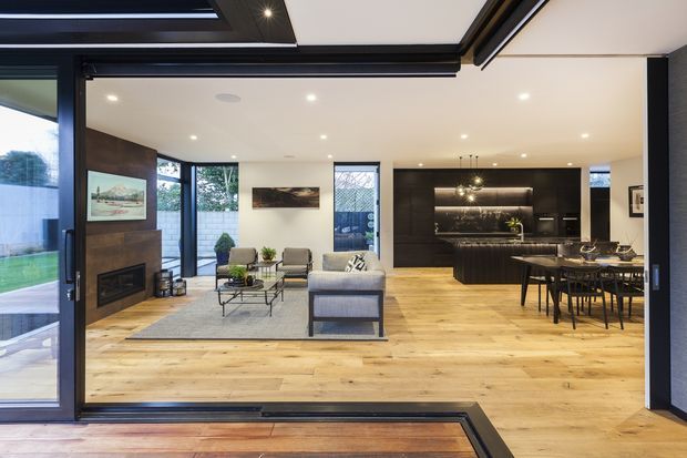 Open-plan contemporary living, dining and kitchen interior with warm timber flooring, dark built-in kitchen cabinetry, a central fireplace feature wall, extensive black-framed glazing, and a partially visible skylight or roof opening. The space is bright, refined, and connected to an outdoor area through large sliding doors.