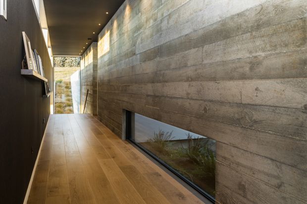 A narrow, elongated architectural hallway with warm timber flooring, a dark wall with recessed display niches on the left, and a long textured concrete feature wall on the right. A low horizontal window is set into the concrete wall, providing a view of planted landscaping, while linear recessed ceiling lights lead toward a glazed opening at the far end. The space has a restrained, contemporary material palette and strong contrast between dark finishes, timber, and raw concrete.