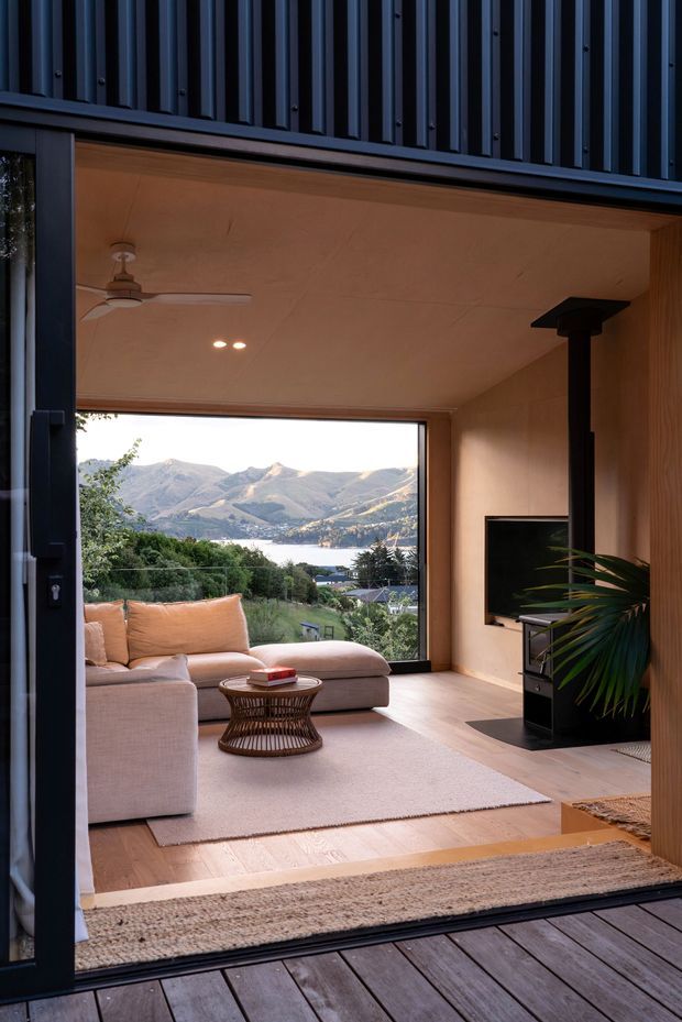 Interior view of a warm, timber-lined living room opening onto a deck through a large glazed sliding door. The room contains a pale sectional sofa, round woven coffee table, television and freestanding black fireplace, with a ceiling fan and recessed lighting. Through the glazing is a clear view of a lake, residential hillside and mountains. Dark vertical exterior cladding frames the opening.
