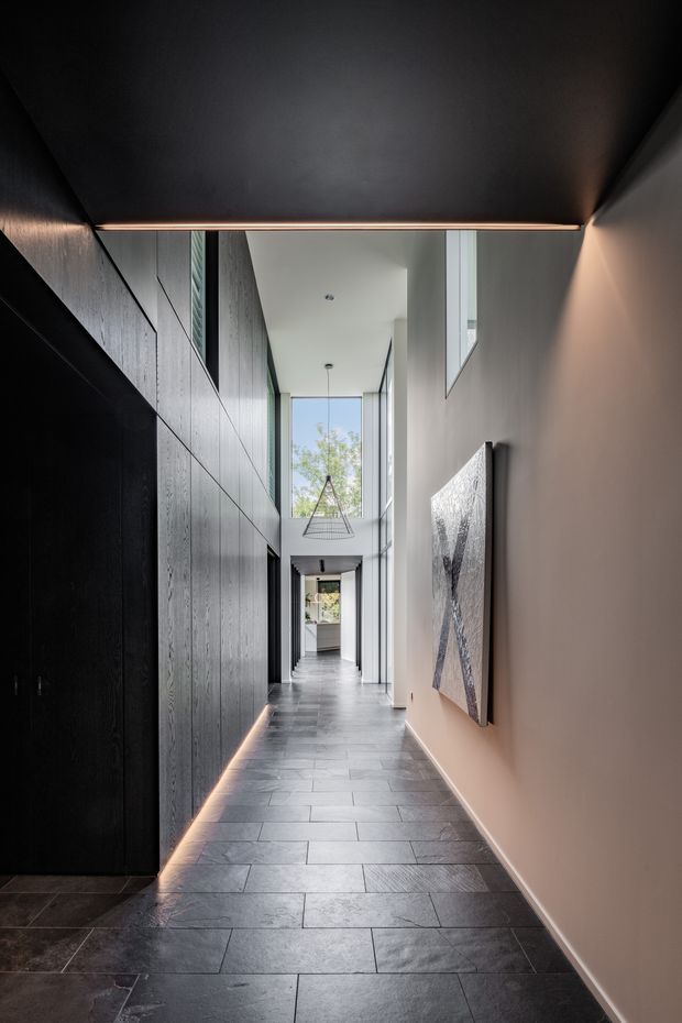 A dramatic, narrow double-height hallway with dark textured built-in wall panelling on the left, a warm neutral plaster wall on the right, and large-format dark grey tiled flooring. Concealed linear lighting traces the base of the dark joinery and a slim ceiling edge, while tall glazing and a suspended sculptural pendant bring daylight into the corridor. Abstract wall art and a glimpse toward another interior space appear at the far end.