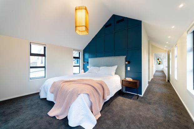 A bright contemporary bedroom with a centrally positioned bed against a dramatic deep-blue paneled feature wall. The room has dark grey carpet, white walls and ceiling, slim black-framed windows, a warm timber pendant light, and a small timber bedside table. A long hallway extends beyond the bedroom, with additional rooms visible in the distance.