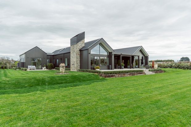 A contemporary single-storey rural residence set within a broad manicured lawn. The home features multiple steep gabled roof forms, dark vertical metal cladding, large glazed gable ends, and a prominent natural-stone chimney. A covered outdoor entertaining area and low stone retaining wall sit along the front elevation.