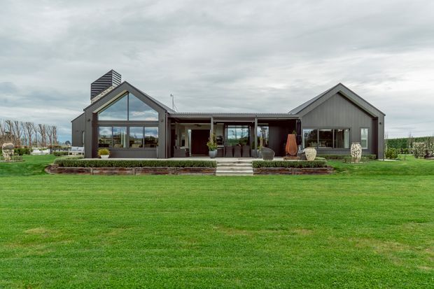 Single-storey contemporary house viewed from the front across an expansive lawn. The building has two pitched gabled wings linked by a recessed central entry and covered terrace, dark vertical cladding, large glazed openings, and a prominent chimney-like feature. Low landscaped retaining beds and clipped hedging frame the facade beneath an overcast sky.