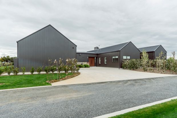 Exterior view of a contemporary single-storey complex composed of several connected gable-roofed volumes. The buildings have dark grey vertical metal cladding, dark roofing, simple rectangular windows and doors, and a broad concrete driveway leading from a gravel road. Low landscaped planting and lawns surround the structures beneath an overcast sky.