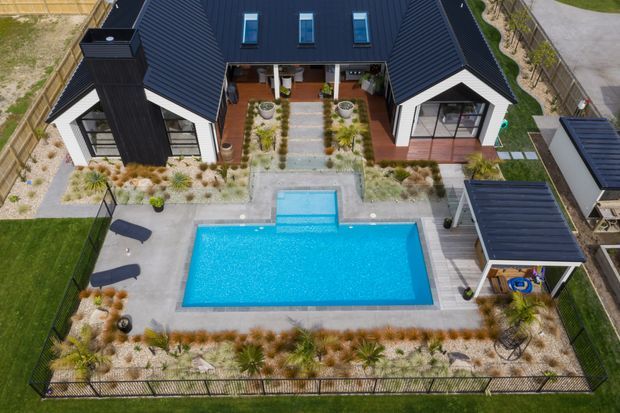 Aerial view of a contemporary dark-clad single-storey home arranged around a landscaped central courtyard, with a bright rectangular swimming pool, paved terrace, garden beds, and a small covered outdoor structure. The house has a complex pitched roof with multiple skylights and white gabled sections, surrounded by manicured drought-tolerant planting and fencing.