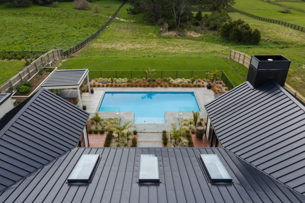Elevated view over a contemporary dark metal-clad roofscape with multiple skylights, enclosing a landscaped swimming pool courtyard and outdoor entertaining area. The property sits within an expansive green rural landscape with fencing and pasture beyond.