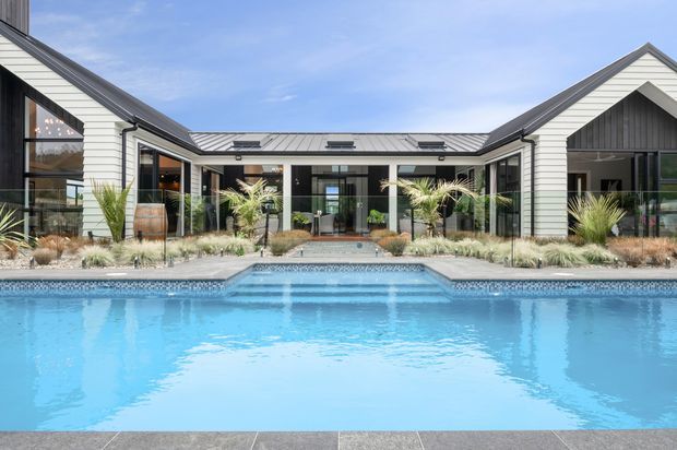 A symmetrical single-storey courtyard-style residence viewed across a large swimming pool. The home has white horizontal cladding, dark metal gabled roofs, extensive black-framed glazing, and a central covered outdoor connection. Gravel landscaping with ornamental grasses and palms surrounds the pool and entry.