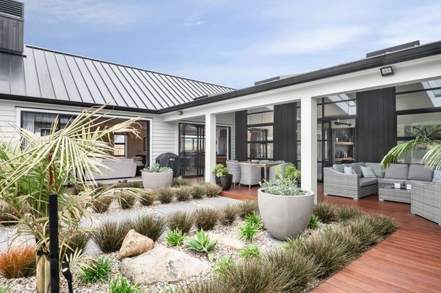 Bright courtyard view of a contemporary single-storey home with a low-pitched metal roof, white exterior walls, dark vertical timber-look panels, and extensive floor-to-ceiling glazing. A timber deck surrounds a landscaped central garden featuring gravel, ornamental grasses, succulents, rocks, large planters, and outdoor wicker seating.