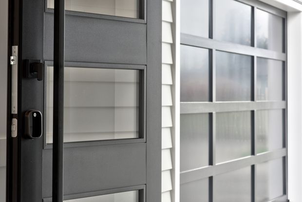 Close-up architectural detail of a dark charcoal entry door with inset rectangular glazing and a modern electronic lock, beside a large grid-framed window with translucent glass and white horizontal weatherboard cladding.