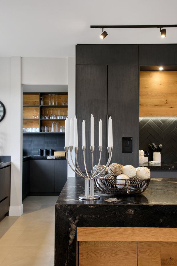 A sophisticated contemporary kitchen viewed toward a dark timber and black joinery wall. The foreground features a large island with a dramatic dark stone benchtop and warm timber cabinetry, while the rear includes open glassware shelving, black lower cabinets, a patterned dark splashback, and a warm timber feature panel illuminated by concealed lighting. Track lighting, a tall sculptural candelabra, and decorative objects add a refined, dramatic finish.