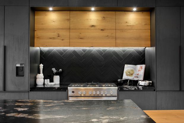 A sophisticated contemporary kitchen viewed straight on, featuring dark matte cabinetry, a large natural-stone island benchtop, a stainless-steel range cooker, and a dramatic black geometric tiled splashback. Warm timber overhead cabinetry with integrated downlights provides contrast and warmth.