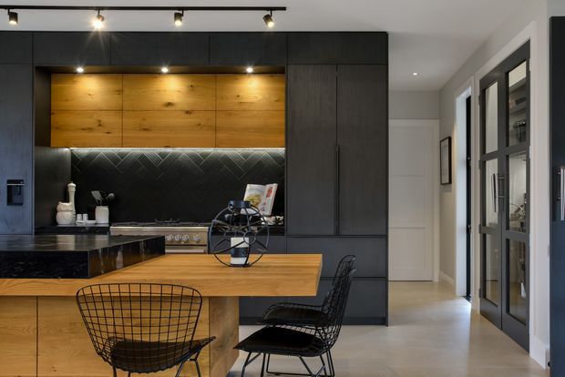 A sophisticated contemporary kitchen with extensive matte dark cabinetry, a warm timber wall niche and island, dark stone benchtops, and a patterned dark splashback. A black wire dining chair and stools sit at the island. Recessed and track lighting illuminate the space, while glazed black-framed internal doors and a hallway extend to the right.