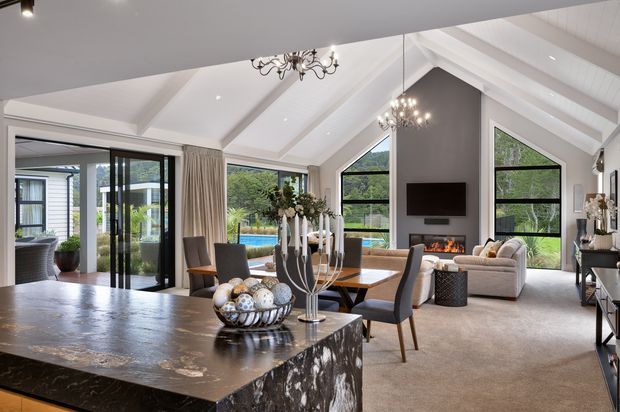Open-plan living and dining area with a kitchen island in the foreground, featuring a dramatic vaulted white ceiling with exposed beams, large black-framed windows and sliding doors, a fireplace wall, carpeted seating area, and views toward an outdoor pool and greenery. The space combines contemporary architecture with refined traditional furnishings and chandeliers.