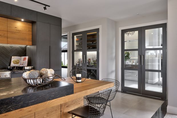 Contemporary kitchen and dining area with dark charcoal cabinetry, a large black stone island, warm timber cabinetry and dining table, patterned dark splashback, and black-framed glazed doors. A glass-fronted drinks or pantry cabinet is visible behind the dining area. The space is well-lit with a refined monochromatic palette softened by natural timber.