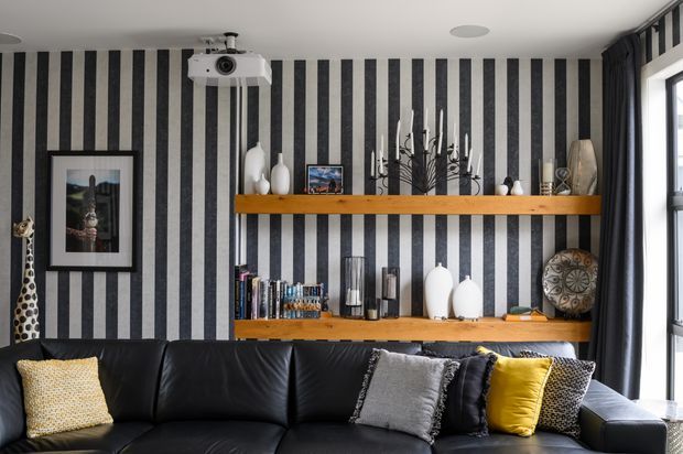 A stylish living room featuring a large black leather sectional sofa, bold black-and-white vertical striped wallpaper, floating timber display shelves with books and decorative objects, framed artwork, a wall-mounted projector, and dark curtains beside a large window. The room has a dramatic monochrome palette with mustard and neutral accent cushions.