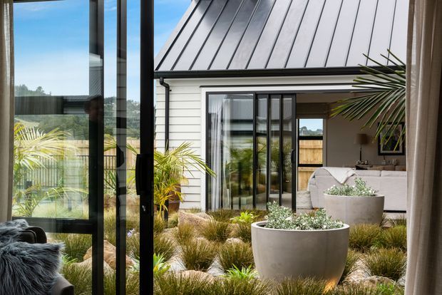View from inside a contemporary home through full-height black-framed glazing toward a landscaped courtyard and a white weatherboard pavilion. The courtyard features ornamental grasses, tropical planting, large pale concrete planters, a timber fence, and a partially visible outdoor seating area. The pavilion has a steep standing-seam metal roof and glazed black-framed doors.