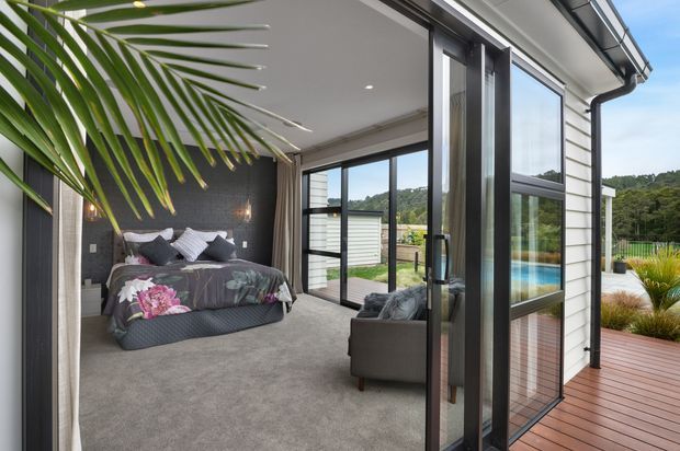 A bright contemporary bedroom opening onto a timber deck through large dark-framed glazed sliding doors. The room has grey carpeting, a dark textured feature wall behind the bed, neutral bedding with floral accents, and a grey armchair. The deck overlooks a landscaped outdoor area with a visible swimming pool, while white horizontal exterior cladding and black-framed glazing define the building envelope.