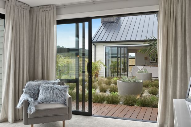 A bright, refined interior seating area viewed toward a landscaped courtyard through expansive black-framed glazed doors. A neutral upholstered armchair with a textured grey throw sits beside full-height beige curtains, while timber decking, ornamental grasses, palms, and a light-clad pavilion with a standing-seam metal roof are visible outside.