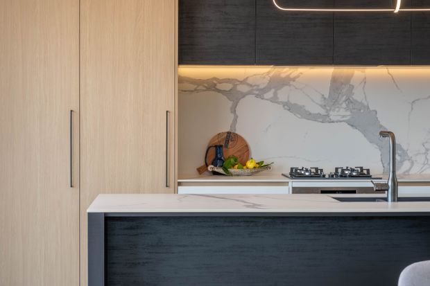 Contemporary kitchen featuring a large island with a dark textured front, pale timber cabinetry, a white marble-look benchtop and full-height splashback, integrated gas cooktop, and a curved brushed-metal tap. Warm concealed under-cabinet lighting and a linear pendant create a refined, high-contrast fit-out.