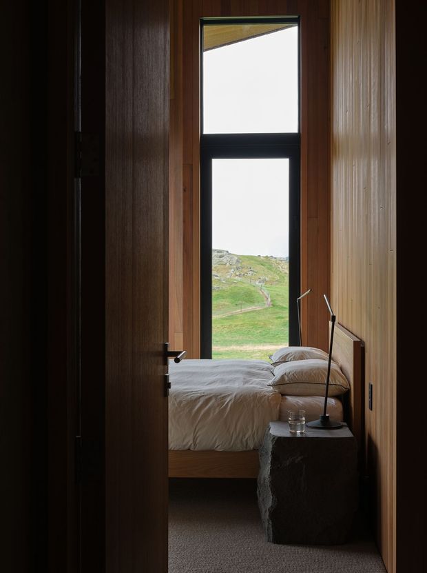 A narrow, moody view into a compact bedroom through a partially open dark timber door. The room features warm vertical timber wall panelling, a low bed with white bedding, a rough-hewn dark stone bedside table, and slim black bedside lamps. A tall black-framed window provides a strong vertical focal point and views of a green rocky hillside outside.