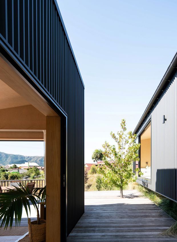 A bright architectural exterior view through a narrow passage between two contemporary buildings. The left structure has dark vertical metal screening and a deep timber-lined opening with a covered outdoor sitting area, while the right structure features light corrugated cladding, dark gutters, and a recessed window. A timber deck walkway leads between the buildings toward landscaped planting and distant hills.