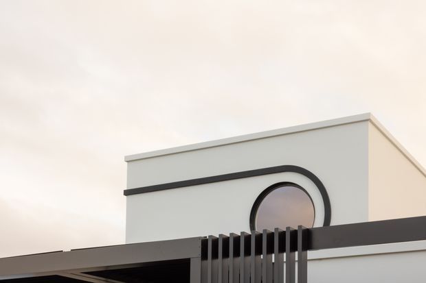 Close-up exterior architectural detail of a contemporary white rendered building, featuring a parapet roofline, a rounded arched window with dark trim, and a dark horizontal canopy or fascia with vertical metal screening in the foreground.