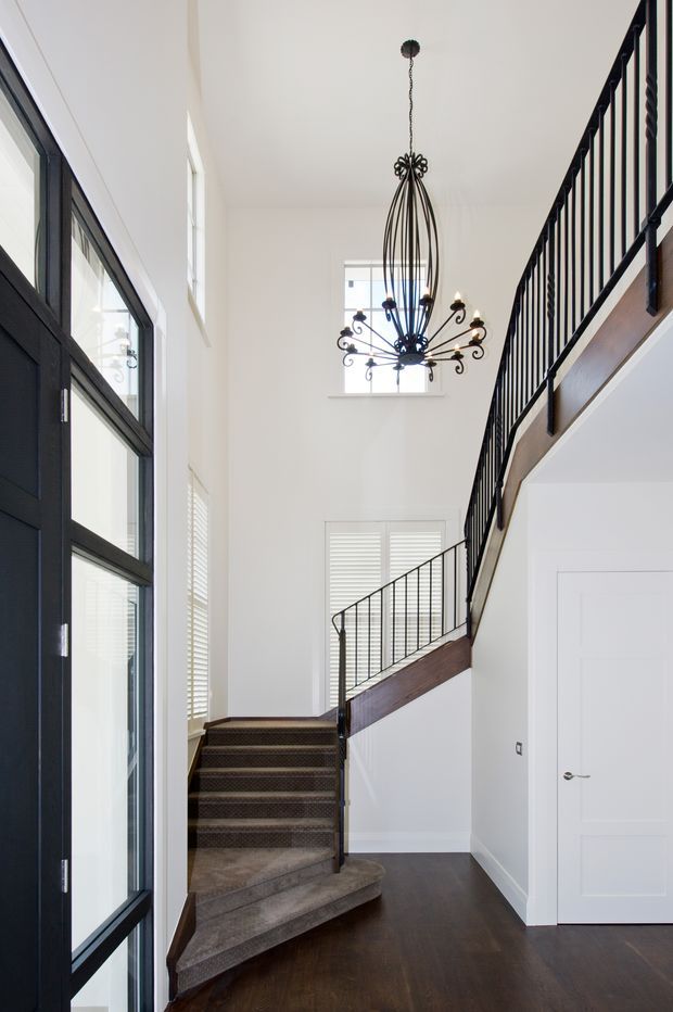 Bright, tall entrance hall with a curved carpeted staircase, dark timber flooring, white walls, black-framed glazing, and a mezzanine-level walkway with black metal balustrades. A decorative black pendant chandelier hangs in the double-height space, and white plantation shutters and doors reinforce the crisp monochrome palette.