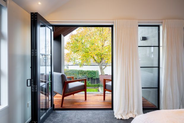 A bright contemporary interior opening onto a timber deck through fully glazed black-framed folding doors. The room has pale walls, recessed lighting, grey carpet, and floor-to-ceiling white curtains. Two timber-framed lounge chairs sit outside on the deck, overlooking a landscaped garden with a mature tree. A portion of a bed is visible in the foreground, indicating a bedroom.