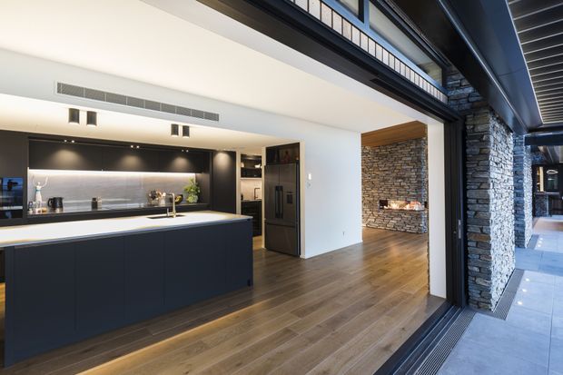 Contemporary open-plan kitchen and living area viewed from a large glazed opening. The kitchen features a dark island and cabinetry, a light benchtop, integrated appliances, and a metallic backsplash. Warm timber flooring leads to a stone-clad fireplace, while dark structural framing and extensive glazing connect the interior to an exterior paved area.