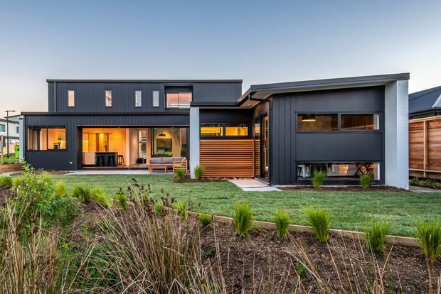 Exterior view of a contemporary multi-volume home with dark vertical cladding, flat and low-pitched rooflines, extensive glazing, warm interior lighting, a timber slatted entry screen, and landscaped native-style planting in the foreground.