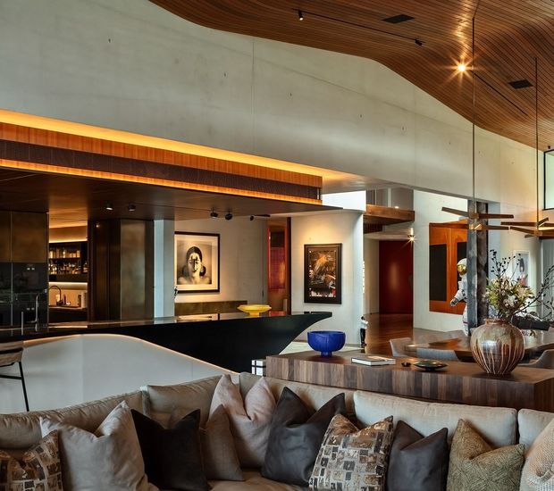 A sophisticated open-plan living and entertaining space with a large sectional sofa in the foreground, a sculptural timber dining or console table, and a dark built-in bar area to the left. The room features exposed concrete walls, extensive warm timber ceiling and joinery, recessed and concealed lighting, large glazed openings, artwork, and curated contemporary furnishings. A partial kitchen or bar zone and circulation spaces are visible beyond the main lounge.