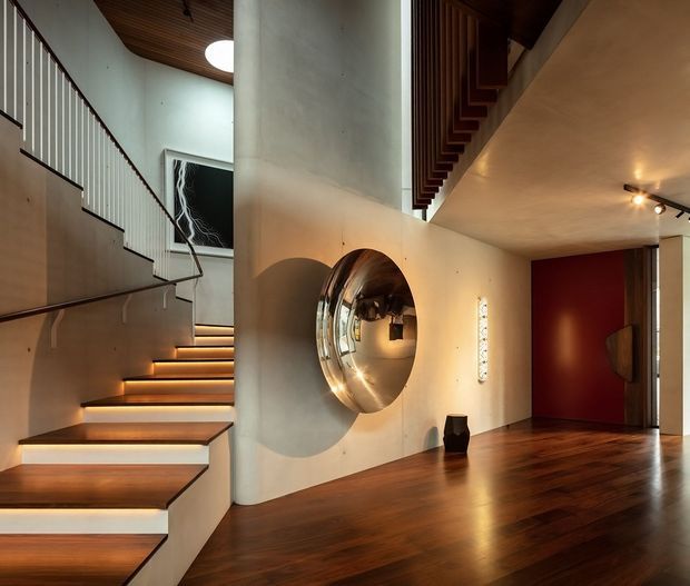 A sophisticated residential entry hall with a sculptural open staircase, warm integrated stair lighting, dark timber flooring, curved white plaster walls, a large circular wall mirror, and a deep red pivot door. Vertical balustrades and timber ceiling details add strong architectural character, while concealed and track lighting create a warm, refined atmosphere.