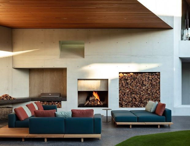 A contemporary open-plan living area with a deep blue modular sectional sofa, red and cream cushions, a low timber coffee table, and a matching chaise. The room features a white rendered or concrete fireplace wall, a built-in open fire, recessed firewood storage, a dark stone or textured hearth bench, and a dramatic angled timber-lined ceiling. A small patch of lawn is visible at the foreground edge.