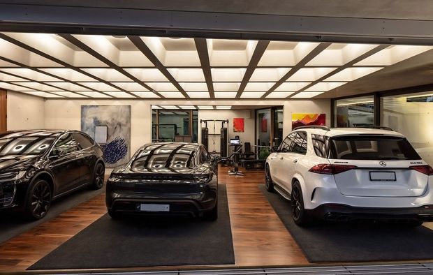 A high-end indoor vehicle showroom or private garage displaying three cars on dark mats over warm timber flooring. The space features a dramatic illuminated grid ceiling with dark structural framing, large glazed openings, artwork, and a gym or exercise area visible toward the rear.