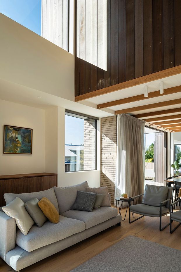 Bright double-height living room with a grey upholstered sofa, armchair, timber flooring, dark vertical timber wall panelling, exposed timber ceiling beams, white-painted brick, large windows and sliding glazed doors opening to an outdoor area. The room has a warm contemporary character with abundant natural light and muted neutral furnishings.