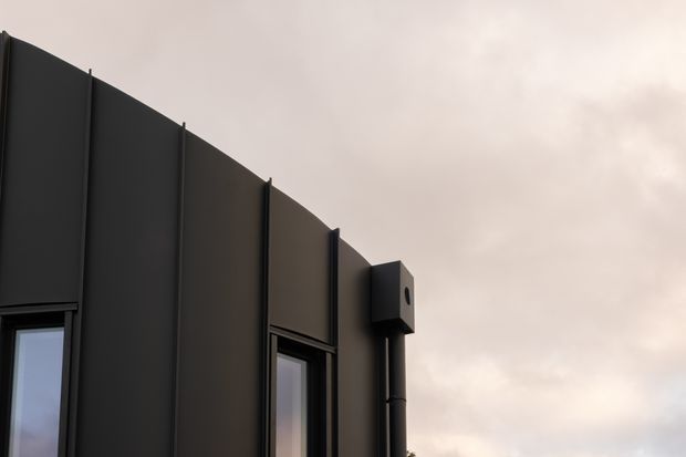 Close-up exterior view of a dark modern building facade with vertically articulated metal cladding panels, narrow black-framed windows, and a projecting box-like architectural detail with a downpipe. The curved or sloping roofline is silhouetted against an overcast sky.