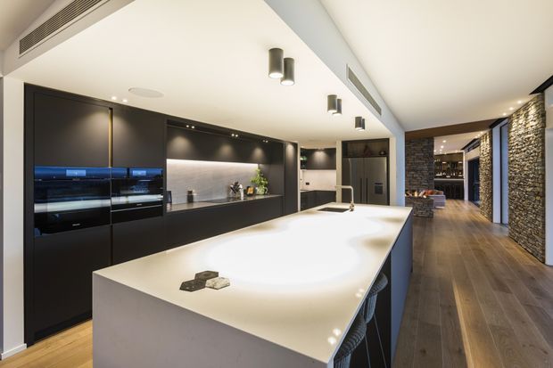 A spacious contemporary kitchen centred on a large white island with an integrated sink and seating. The room features extensive matte-black cabinetry, integrated appliances, a dark textured backsplash, recessed and pendant-style lighting, timber flooring, and a stone-clad feature wall extending toward an adjoining wine or display area.