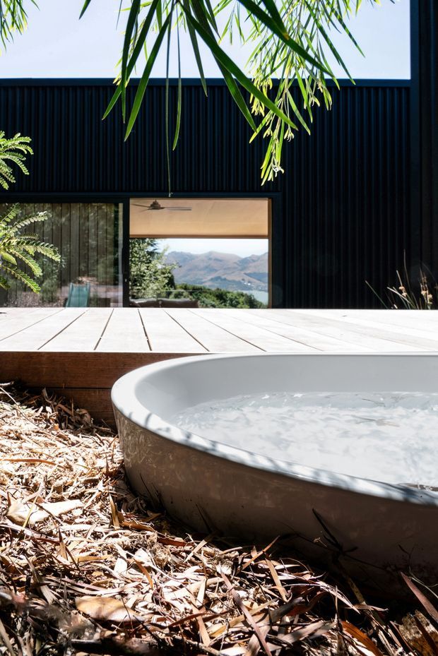 A contemporary black-clad house viewed from an outdoor deck, with a freestanding oval bathtub in the foreground surrounded by leaf litter and planting. Large glazed openings reveal a warm interior and expansive mountainous scenery beyond. The composition has a strong indoor-outdoor connection and a refined, nature-integrated feel.