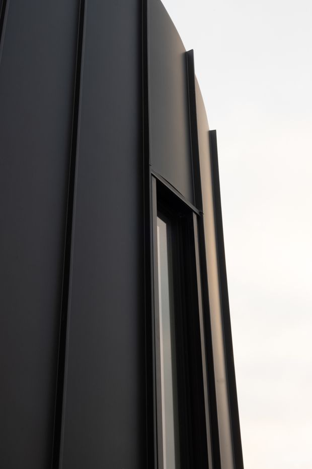 Close-up, low-angle view of a tall contemporary building facade with dark vertical cladding or metal fins framing a narrow window opening. The composition emphasizes sleek vertical lines, deep reveals, sharp geometry, and a restrained black and charcoal palette against a bright sky.
