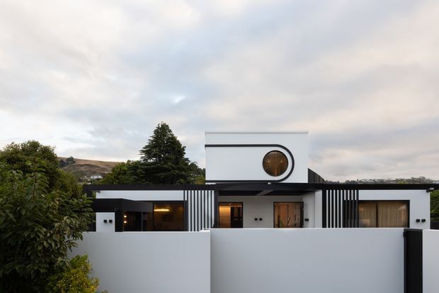 Front exterior view of a contemporary low-rise residence with a crisp white rendered facade, strong horizontal rooflines, black detailing, vertical slatted screens, and a distinctive circular window. A solid white boundary wall and gate frame the property, with landscaped planting and distant hills visible around it.