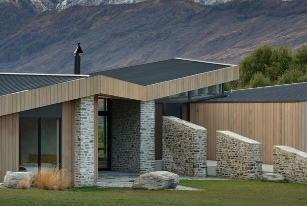 A low-slung contemporary mountain house with dark standing-seam metal roofs, vertical timber cladding, extensive glazing, and prominent rough stone walls. The building is set against a dramatic alpine mountain backdrop, with a landscaped lawn, ornamental grasses, boulders, and a sculptural stone boundary wall in the foreground.