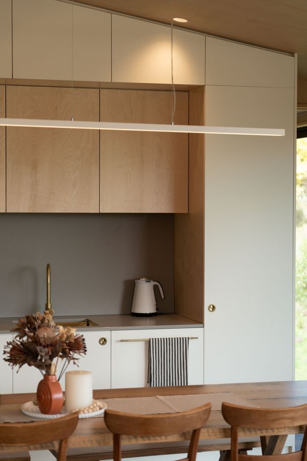A warm, minimalist kitchen and dining area with integrated cabinetry. Natural timber overhead cupboards and backsplash surround matte off-white cabinetry, a dark grey benchtop and splashback, brass-toned tapware and hardware, and a slim linear pendant light. A timber dining table and wooden chairs occupy the foreground, with a window bringing in daylight at right.