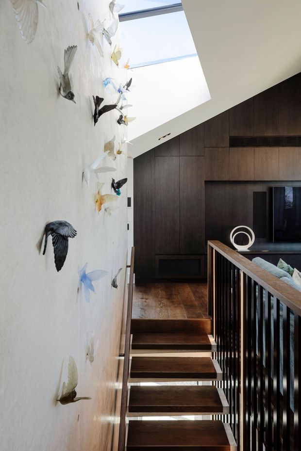 A contemporary interior stairway with floating dark timber treads, slim black vertical balustrades, and dark timber wall panelling. A large skylight brightens the stairwell, while a textured pale wall is decorated with sculptural bird forms. The stair landing opens toward a media or living area with a television and low joinery.
