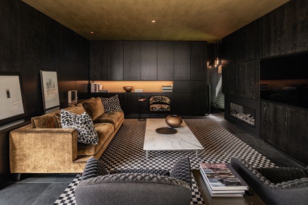 A dark, sophisticated living room with a low ceiling and extensive charcoal-black wall panelling and built-in cabinetry. A brown velvet sofa, charcoal quilted armchairs, patterned black-and-white rug, marble-look coffee table, built-in fireplace, recessed television and warm accent lighting create a luxurious, moody interior.